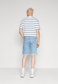 Bermudas de mezclilla azul claro con dobladillos enrollados, combinadas con una camiseta blanca a rayas. Modelo de espaldas, usando calcetas blancas y zapatillas deportivas.