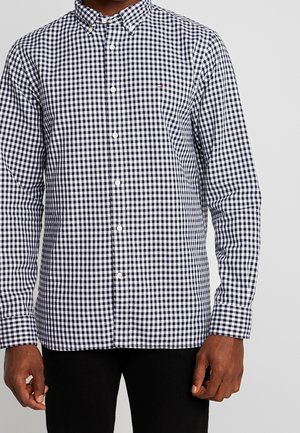 Homme portant une chemise à carreaux noir et blanc à manches longues avec un petit logo sur la poitrine, associée à un pantalon noir.