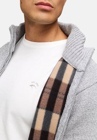 Grå strikjakke med en fremtrædende krave, lagd ovenpå en hvid skjorte med et diskret logo, suppleret med et mønstret tørklæde i beige, brun og grå.