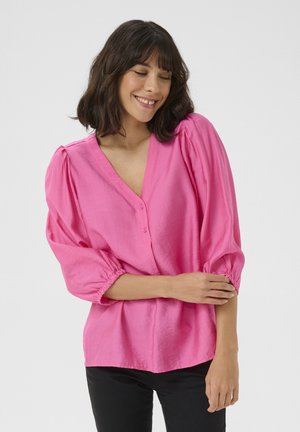 Blouse rose, légère, avec un col en V, des détails de bouton et des manches bouffantes. Elle présente des poignets froncés et une coupe ample, fabriquée à partir d'un tissu doux.