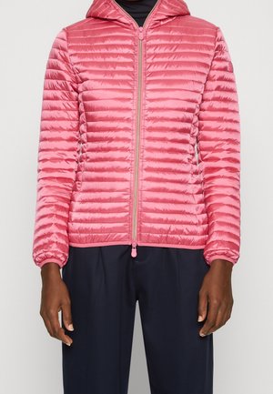 Personne portant une veste matelassée rose à capuche avec une fermeture éclair à l'avant, associée à un pantalon bleu marine foncé sur un fond clair uni.