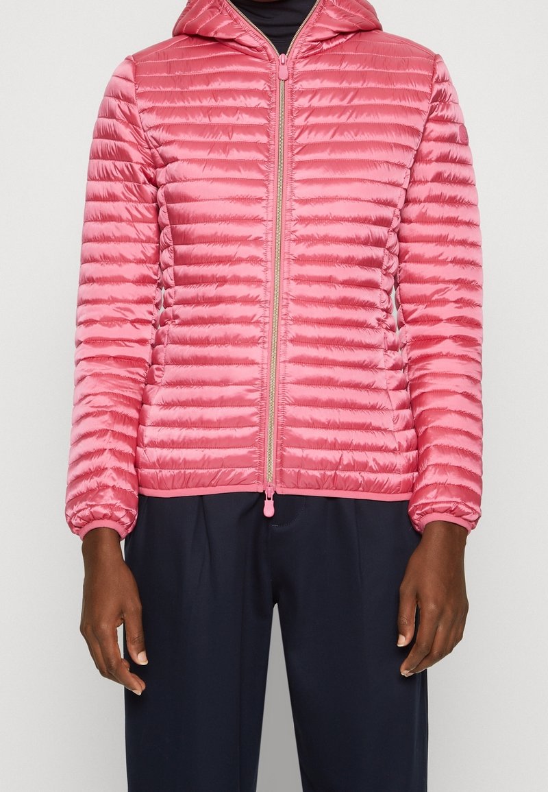 Personne portant une veste matelassée rose à capuche avec une fermeture éclair à l'avant, associée à un pantalon bleu marine foncé sur un fond clair uni.