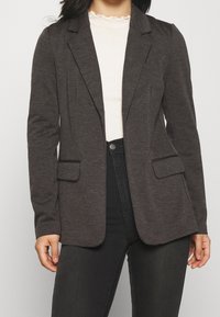 Grijze blazer van zacht materiaal, met een getailleerde pasvorm, notch-revers, twee voorzakken en een gladde textuur. Draagt over een geribd topje.