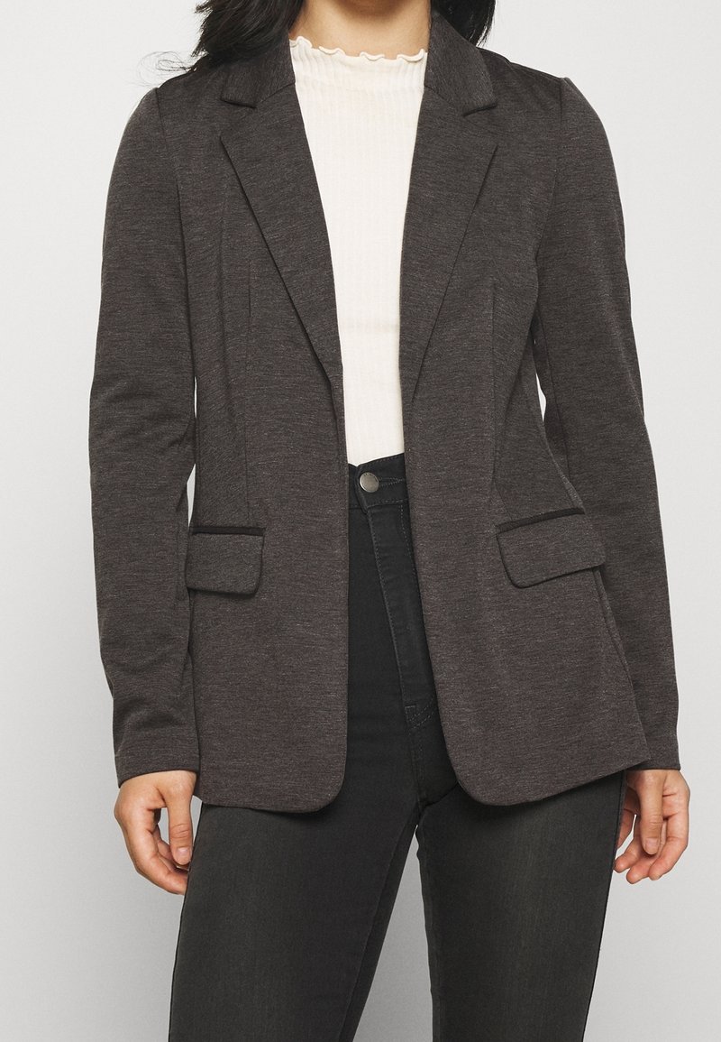 Grijze blazer van zacht materiaal, met een getailleerde pasvorm, notch-revers, twee voorzakken en een gladde textuur. Draagt over een geribd topje.