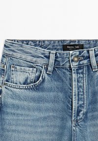 Světle modré džíny z denimu s vysokým pasem, s přední kapsou, kovovým knoflíkem a ozdobnými cvoky podél boční strany. Viditelná značka značky.