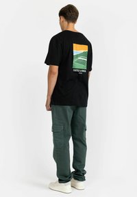 Black cotton t-shirt featuring a colorful graphic on the back with landscape and tennis court designs. Paired with green cargo pants.