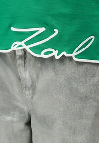 Πράσινο ύφασμα με λευκό χειρόγραφο κείμενο "Karl" κεντημένο πάνω σε ξεβαμμένο γκρίζο τζην.