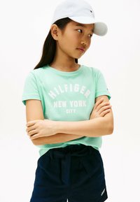 Camiseta de manga corta verde menta con "HILFIGER NEW YORK CITY" impreso en blanco, combinada con pantalones cortos azul marino oscuros con un cinturón de lazo.