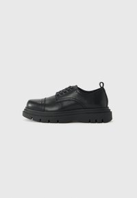 Chaussure en cuir noir avec lacets, semelle épaisse et languette au talon, vue de côté sur fond clair uni.