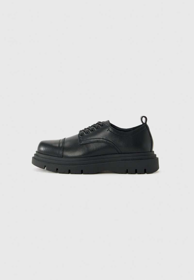 Chaussure en cuir noir avec lacets, semelle épaisse et languette au talon, vue de côté sur fond clair uni.