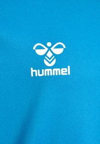 Camiseta de algodón azul con un logo blanco que presenta un diseño de abeja estilizado y la palabra "hummel" debajo. Textura suave.