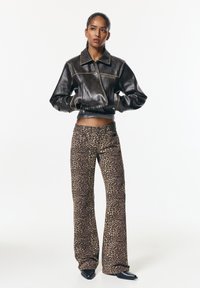 Femeie stă cu mâinile în buzunare, purtând o jachetă din piele maro închis, scurtă, și pantaloni evazați cu model leopard, completând look-ul cu cizme negre cu vârf ascuțit.