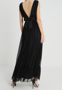 Černé maxi šaty s hlubokým V výstřihem vzadu, přiléhavým korzetem, splývavou skladanou sukní a volánovým lemem, zvýrazněné velkou mašlí v pase.