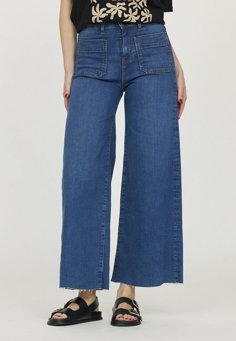 Pantalons en denim bleu à jambes larges avec une taille haute, dotés de deux poches avant et de détails à ourlet effiloché. Portés avec des sandales noires.