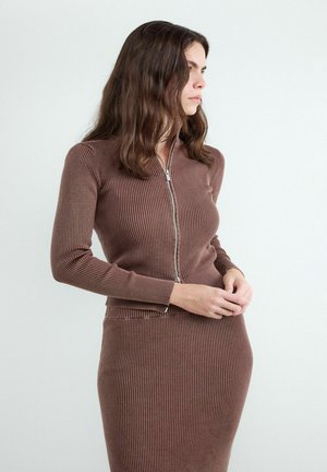 Vrouw met golvend bruin haar die een aangesloten geribde bruine trui met rits en bijpassende rok draagt, kijkt naar rechts met gevouwen handen.