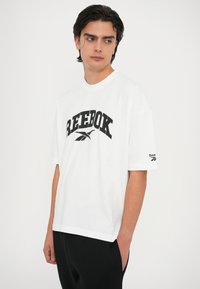 Bijela oversized majica s crnim "Reebok" logotipom, kratkim rukavima, okruglim izrezom i malim logom na rukavu. Pamuk.