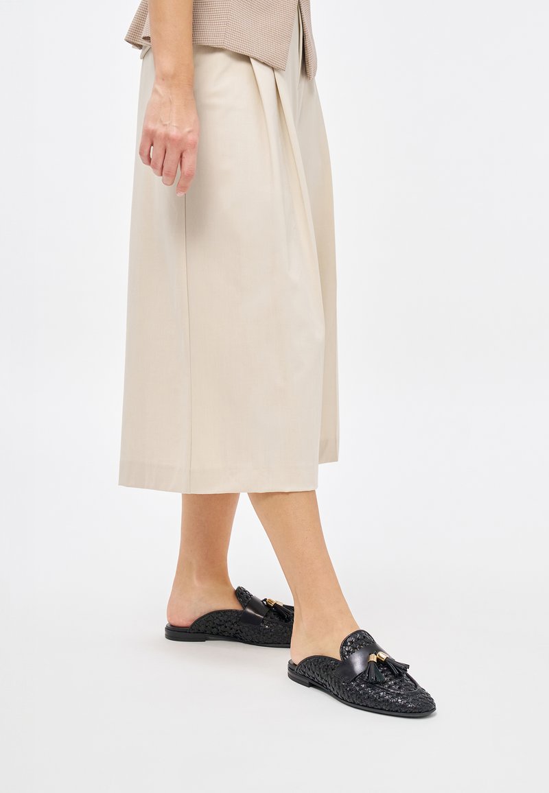 Scarpe slip-on nere intrecciate con dettaglio decorativo a nappina, abbinate a pantaloni beige a gamba larga e una blusa in tonalità marrone chiaro con texture.