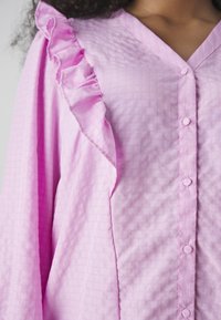 Blusa rosa con un collo a volant, morbida al tatto e con apertura frontale a bottoni. Il tessuto presenta un leggero motivo a quadri su tutta la superfice.