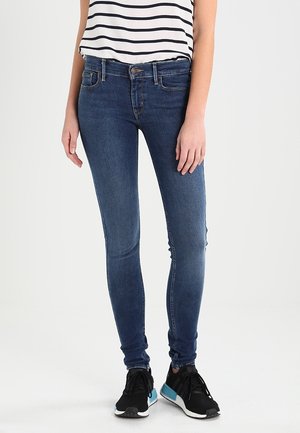 Una persona está de pie con jeans ajustados de color azul oscuro y una camiseta a rayas, llevando zapatillas negras. El fondo es liso y de color claro.