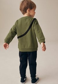 Grøn sweatshirt med sort diagonal stribe, blødt materiale, afslappet pasform, parret med sorte bukser og sorte sneakers. Barn set fra ryggen.