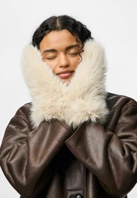 Brązowa skórzana kurtka z szerokim, miękkim kołnierzem w kolorze ecru. Twarz modela częściowo zasłonięta, oczy zamknięte, ręce trzymają kołnierz.