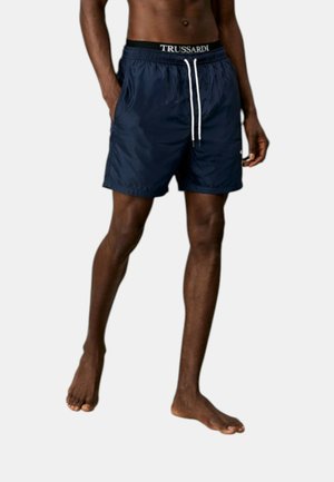 Modello maschile che indossa pantaloncini da bagno blu navy con coulisse bianche e fascia con logo Trussardi, in piedi a piedi nudi su uno sfondo semplice.
