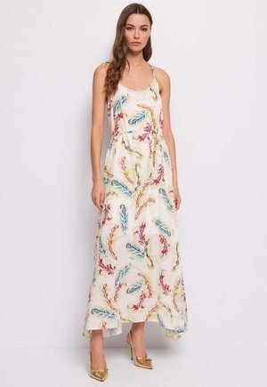 Donna in piedi che indossa un abito lungo bianco senza maniche con motivi di piume colorate e scarpe dorate con tacco alto, su uno sfondo semplice.