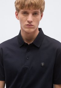 Tengerészkék galléros póló sima anyagból, klasszikus gallérral, háromgombos patenttal és logóval a bal mellrészen.