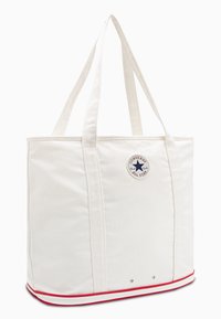 Bijela platnena torba s crvenim prugama, okrugla oznaka s logotipom i dvije čvrste ručke. Ima prostran pravokutni oblik.