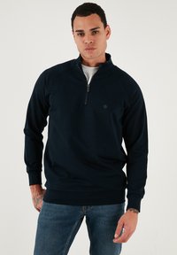 Sötét tengerészkék pulóver, negyed zipzárral ellátott gallérral, hosszú raglán ujjakkal, bordázott mandzsettákkal és egy kis logóval a mellkasán. Simaság jellemzi.