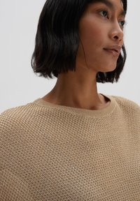 Beige gestrickter Pullover mit einem strukturierten Muster, rundem Ausschnitt und lockerer Passform. Leichter Stoff fällt sanft über die Schultern.