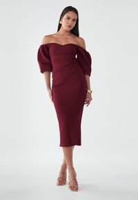 Abito midi bordeaux con spalle scoperte, maniche a palloncino e silhouette aderente. Tessuto morbido e dettagli drappeggiati lungo il corpo.