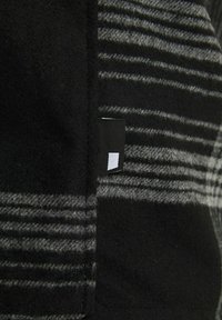 Gros plan d’un tissu en laine rayé noir et gris avec une petite étiquette noire comportant un carré blanc partiel cousu dans la couture.