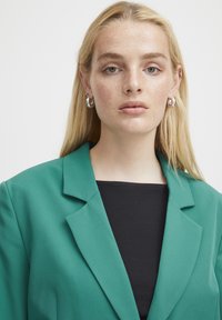 Blazer verde con un diseño estructurado y solapas de muesca, combinado con una blusa negra de canalé, y aretes de plata en forma de aro. Textura suave.