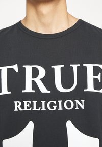 T-shirt noir en coton, portant un large texte blanc qui dit "TRUE RELIGION" en lettres grasses. Encolure ronde et manches courtes.