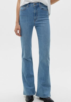 Frau trägt hellblaue High-Waist-Jeans mit Bootcut-Beinen, kombiniert mit schwarzen spitzen Stiefeln und einem weißen, hineingesteckten Hemd.