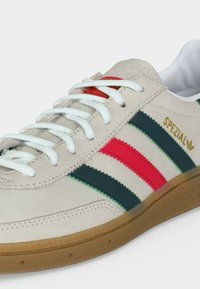 Baskets Adidas Spezial beiges avec lacets blancs, bandes rouges et vert foncé, logo doré et semelle en caoutchouc marron texturée.