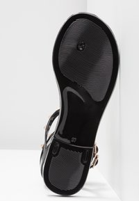 Semelle de sandale noire avec une finition en caoutchouc texturé, présentant un emblème circulaire et une indication de taille. Comprend des accents métalliques et un aspect brillant.