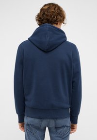 Marineblauer Hoodie mit Kapuze und Kordelzug, der eine weiche Textur und gerippte Bündchen aufweist, getragen zu hellblauen Jeans.