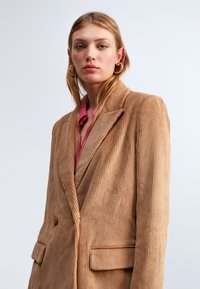 Blazer in corduroy color marrone chiaro con ampi revers, due tasche frontali e texture a coste verticali. Indossato sopra una camicia rosa.