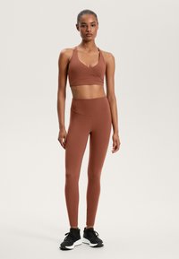 Brown sports bra with wrap design and thin straps, paired with high-waisted brown leggings. Black athletic shoes complete the outfit.