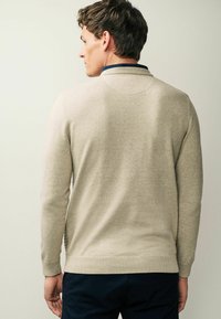 Pull en tricot beige avec un col et des poignets côtelés, présentant un motif texturé sur le côté, porté sur un pantalon sombre.