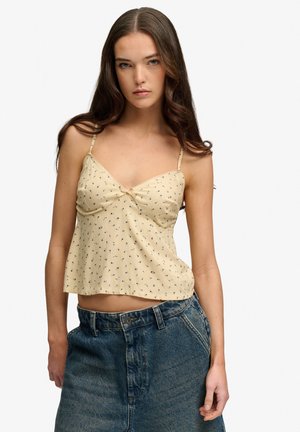 Ung kvinde med langt brunt hår iført en beige blomsterprintet top med spaghetti-stropper og højtaljede blå denimjeans.