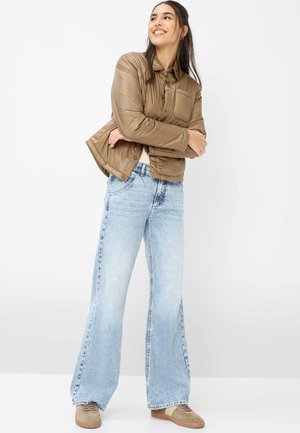 Kvinde smilende, iført brun quiltet jakke, lyseblå vidde bukser og beige sneakers, stående med armene krydsede på hvid baggrund.