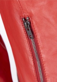 Červená kožená bunda s metalickým zipem, texturovaným povrchem a kontrastním prošíváním podél zipu.