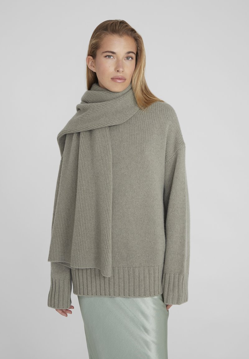 Lichtgrijze gebreide trui met een geribbelde halslijn en manchetten, gecombineerd met een bijpassende geribbelde sjaal die om de nek is gedrapeerd. Gladde, zijdezachte rok zichtbaar.