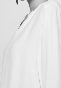 Persona che indossa una blusa bianca larga con scollatura a V e maniche lunghe a sbuffo, mostrata dalle spalle fino a metà torace.
