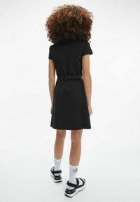 Černé šaty s krátkým rukávem a páskem v oblasti pasu, texturovaná látka, tvar A-line a přiléhavý střih. Kombinováno s bílými ponožkami a černými sandály.