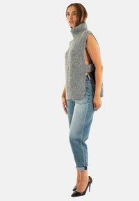 Donna che indossa un maglione a collo alto grigio senza maniche, jeans blu arrotolati e tacchi alti neri, in piedi contro uno sfondo bianco.