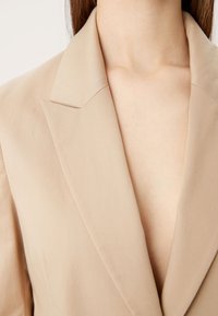 Beige blazer met notch revers, gemaakt van een gladde stof. Heeft een diepe V-hals en een getailleerde pasvorm die de gestructureerde stijl benadrukt.